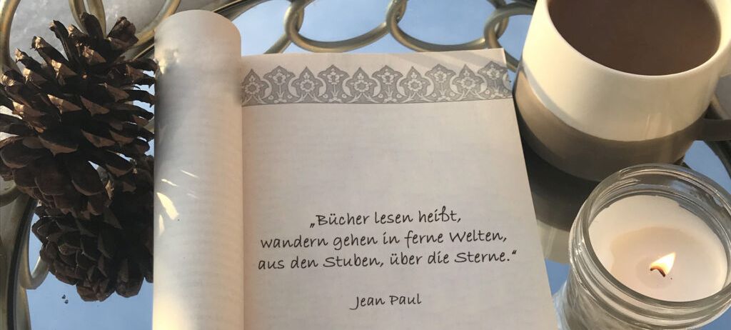 Foto eines aufgeschlagenen Buches mit dem Zitat "Bücher lesen heißt, wandern gehen in ferne Welten, aus den Stuben, über die Sterne. Jean Paul" Daneben eine Tasse, eine Kerze und ein Zapfen