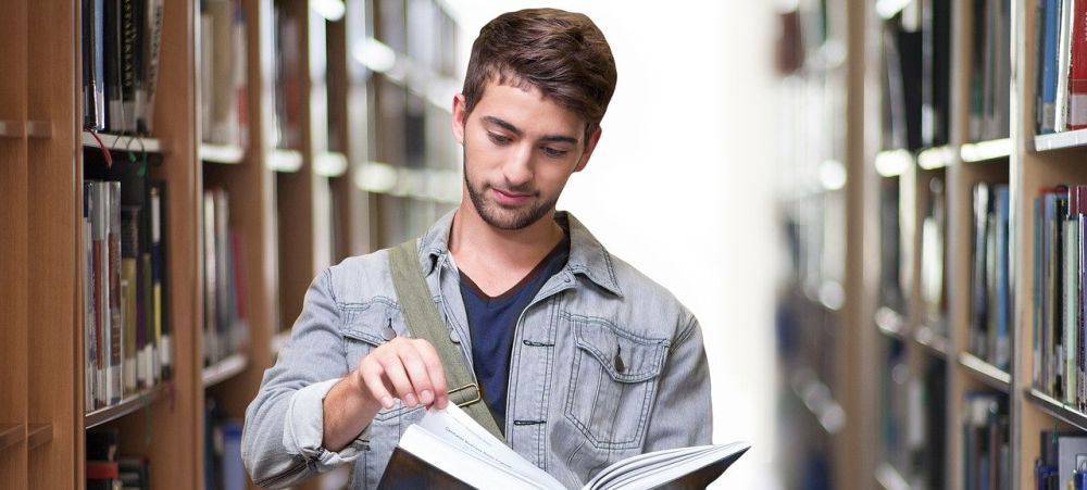 Foto eines jungen Mannes zwischen Bücherregalen, der in einem Buch blättert