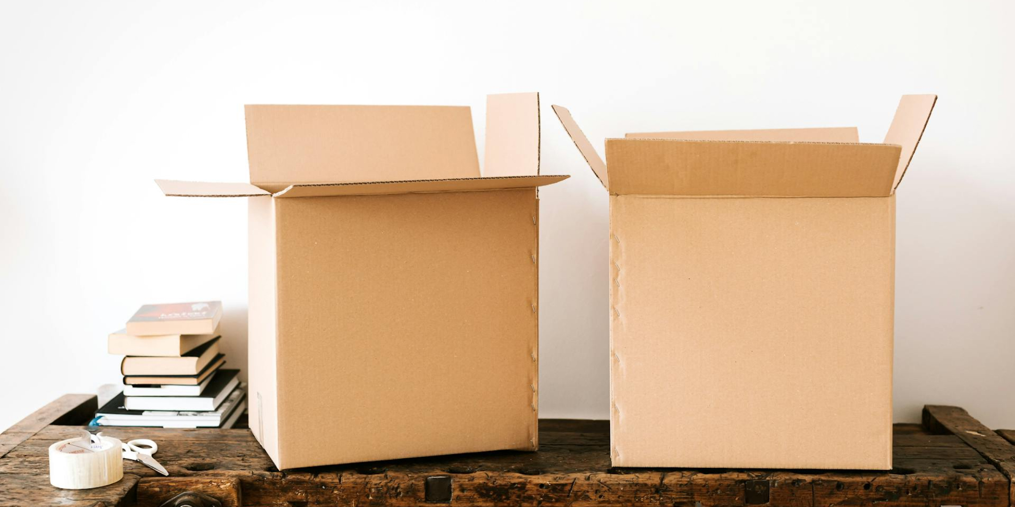 2 Umzugskartons stehen auf einer Holzwerkbank. Links sind Bücher, Schere und Klebeband abgelegt